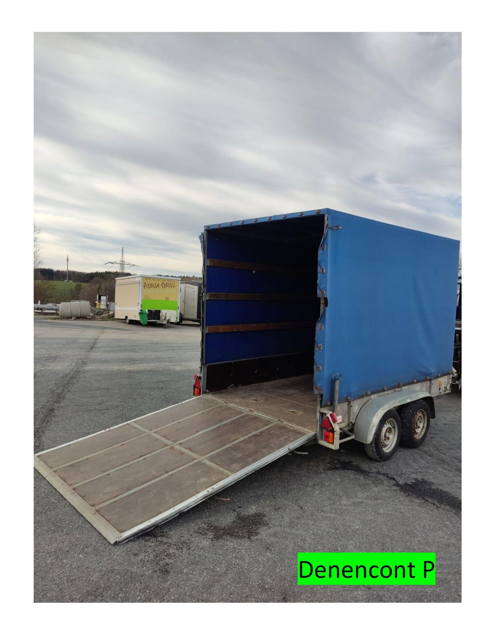 Blauer Anhänger mit offener Rampe auf einem gepflasterten Grundstück bei bewölktem Himmel.