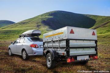 Silbernes Auto, das einen kleinen Anhänger auf einer Grasebene mit Hügeln im Hintergrund zieht.