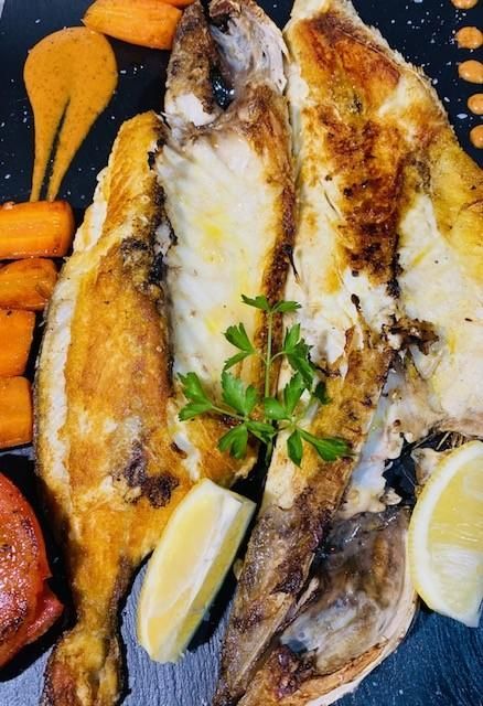 Pescado a la parrilla con zanahorias asadas, salsa y limón en un plato negro.