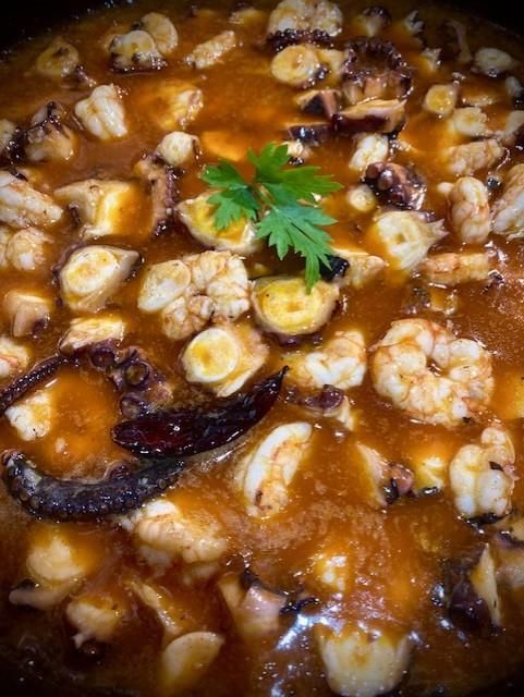 Vista de cerca de un guiso de mariscos en salsa de color marrón anaranjado; incluye camarones, pulpo y chiles secos.