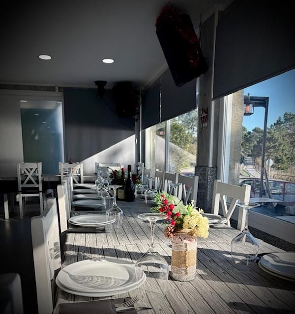 Comedor de restaurante con largas mesas dispuestas con platos, vasos y flores cerca de una ventana que da a una calle.