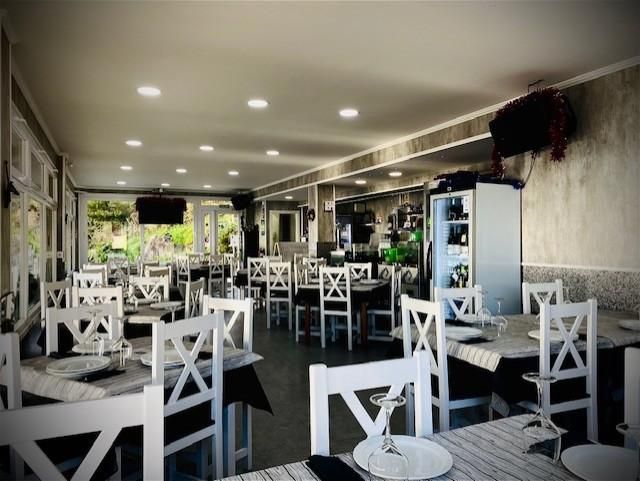 Interior de restaurante con sillas blancas, mesas con platos y vasos. Ventanas a la izquierda.