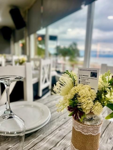 Mesa de restaurante con flores, platos y copa de vino, con vista al mar a través de una ventana.