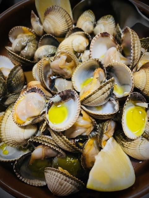 Almejas en concha con aceite amarillo en un recipiente marrón, servidas con gajos de limón.