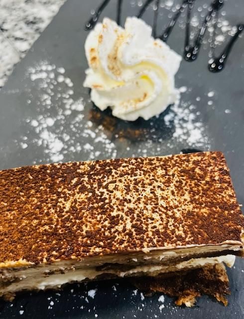 Postre de tiramisú con crema batida y chocolate lloviznado sobre un plato negro, espolvoreado con azúcar glas.