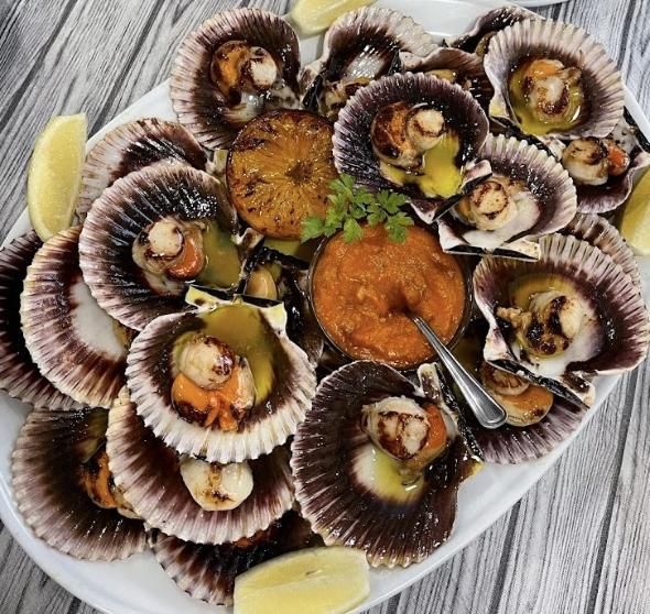 Vieiras a la parrilla con concha en bandeja con salsa y rodajas de limón.