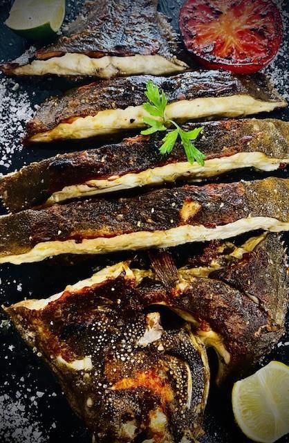 Filetes de pescado fritos con tomate carbonizado y gajos de lima sobre una superficie oscura, adornados con perejil.