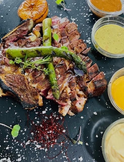 Filete de ternera a la parrilla cortado en rodajas con espárragos y salsas sobre un plato de pizarra oscura.