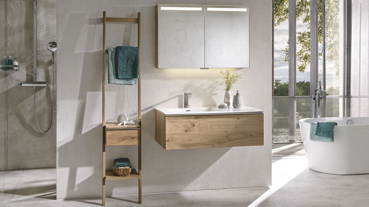 Modernes Badezimmer mit Waschtisch aus Holz, passendem Leiterregal und beleuchtetem Spiegelschrank vor einer strukturierten Steinwand.