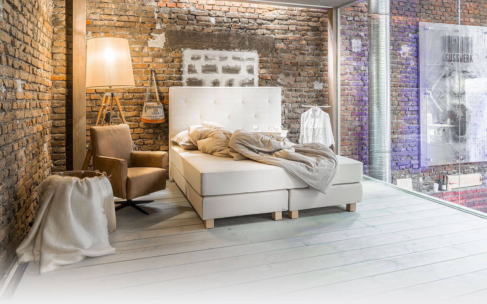 Ein weißes Bett mit gepolstertem Kopfteil, ein beiger Stuhl und eine Stehlampe in einem Zimmer mit rustikalen Backsteinwänden.