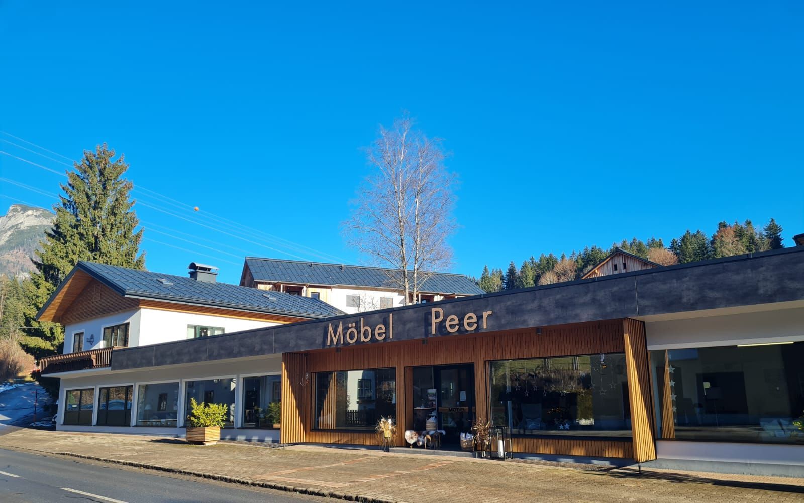 Ein modernes, eingeschossiges Gebäude mit Holzfassade und großen Glasfenstern vor der Kulisse von Bäumen und Bergen.