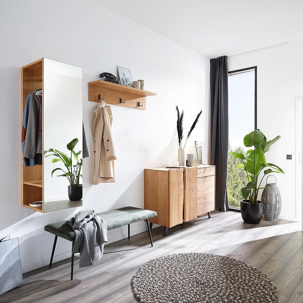 Ein heller Eingangsbereich mit einem Holzschrank, Spiegel, Garderobe, grüner Bank und zwei großen Topfpflanzen auf Holzboden.