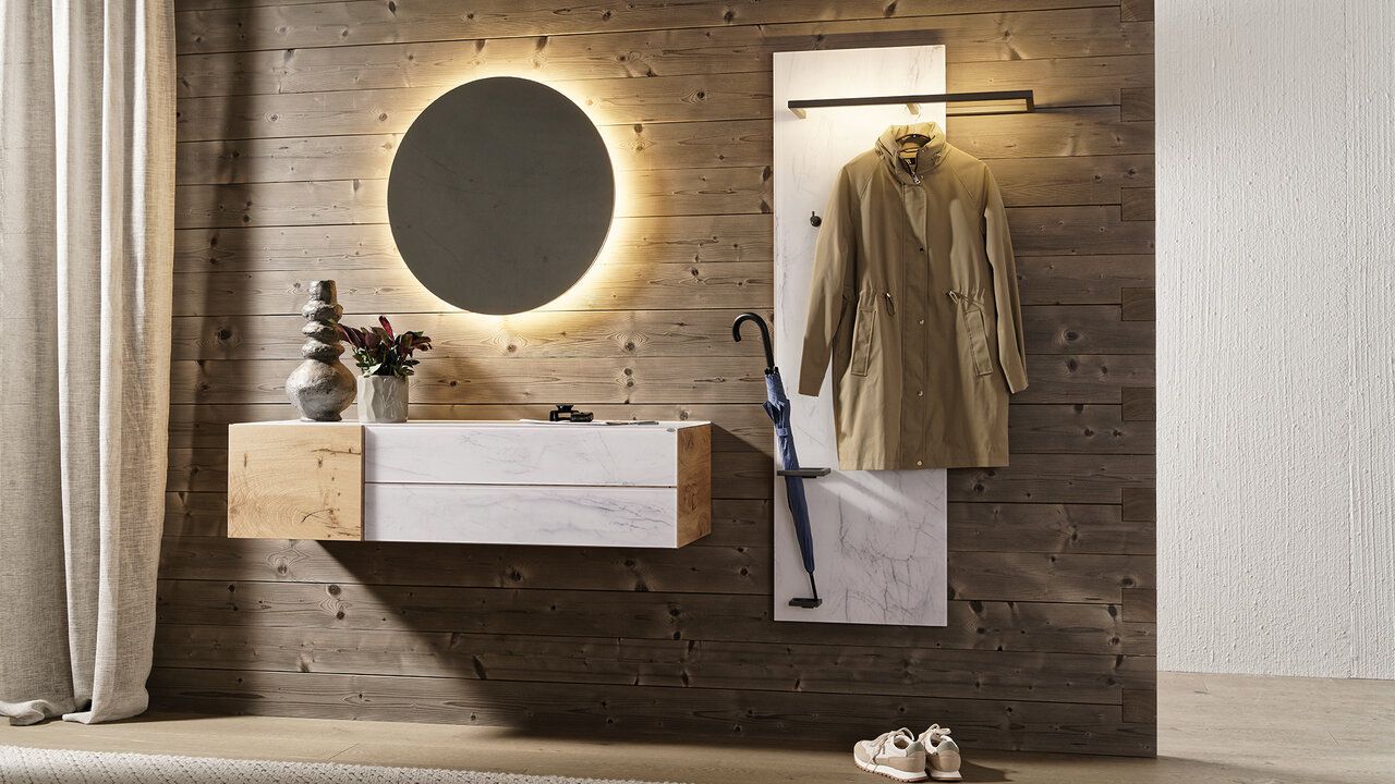 Ein minimalistischer Eingangsbereich aus Holz mit einer schwebenden weißen Konsole, einem hinterleuchteten runden Spiegel und einer Kleiderstange mit Mantel.