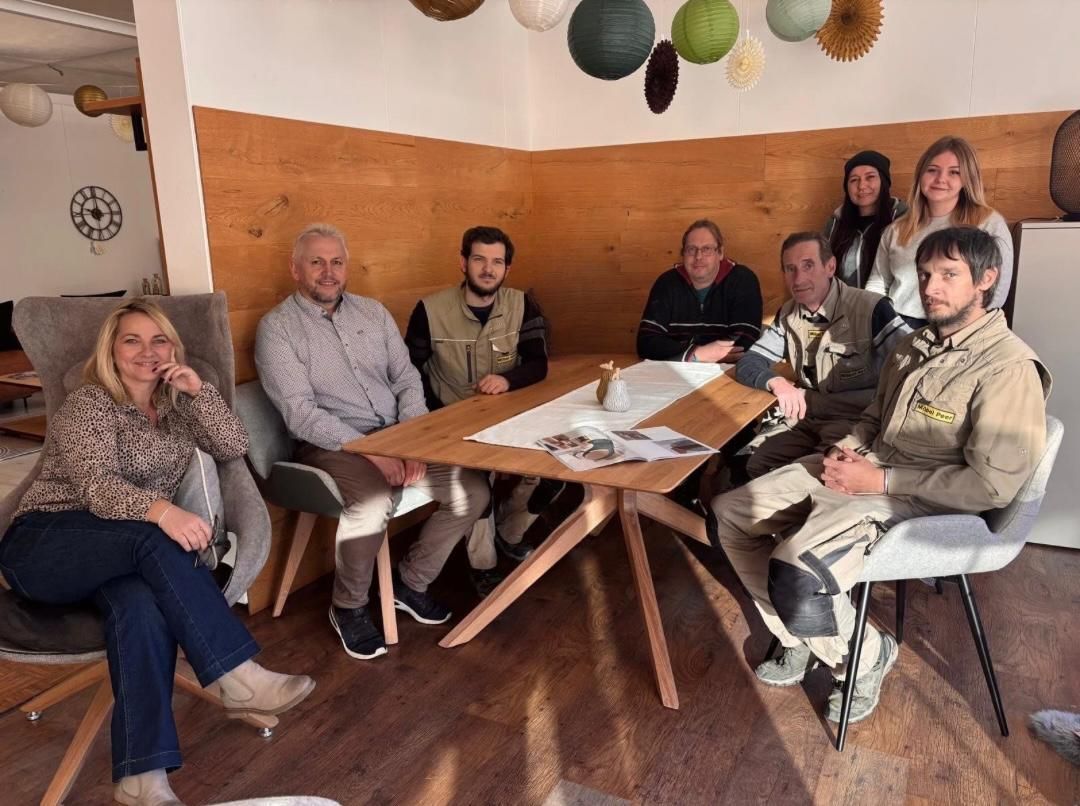 Eine Gruppe sitzt in einem gemütlichen Raum um einen Holztisch und blickt in die Kamera. Auf dem Tisch liegen Papiere und stehen Getränke.