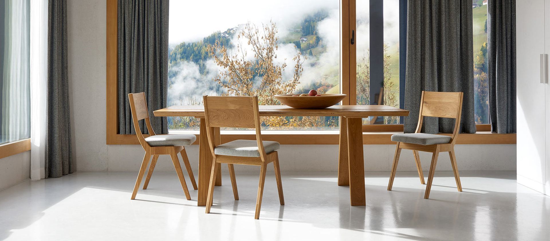 Ein Esstisch mit drei Holzstühlen in einem hellen Zimmer mit großen Fenstern, die den Blick auf eine nebelverhangene Berglandschaft freigeben.