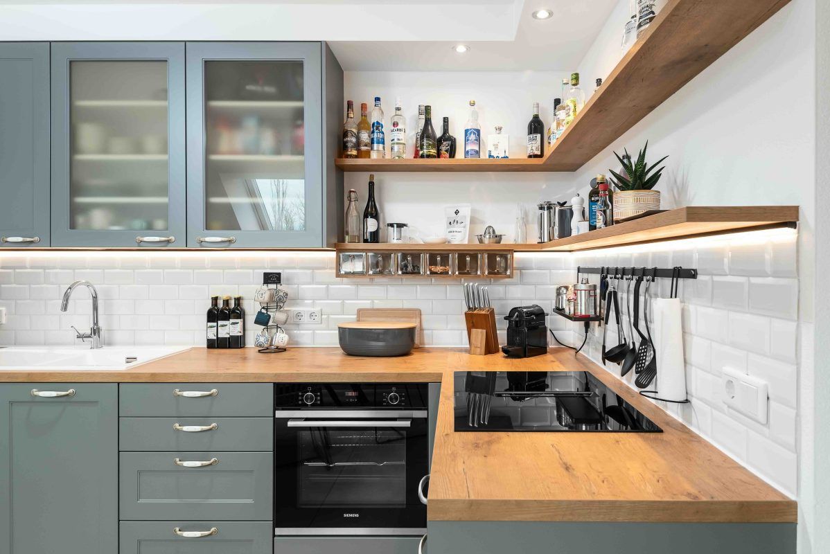 Moderne L-förmige Küche mit grauen Schränken, hellen Holzarbeitsplatten, weißen Metrofliesen als Spritzschutz und offenen Regalen.