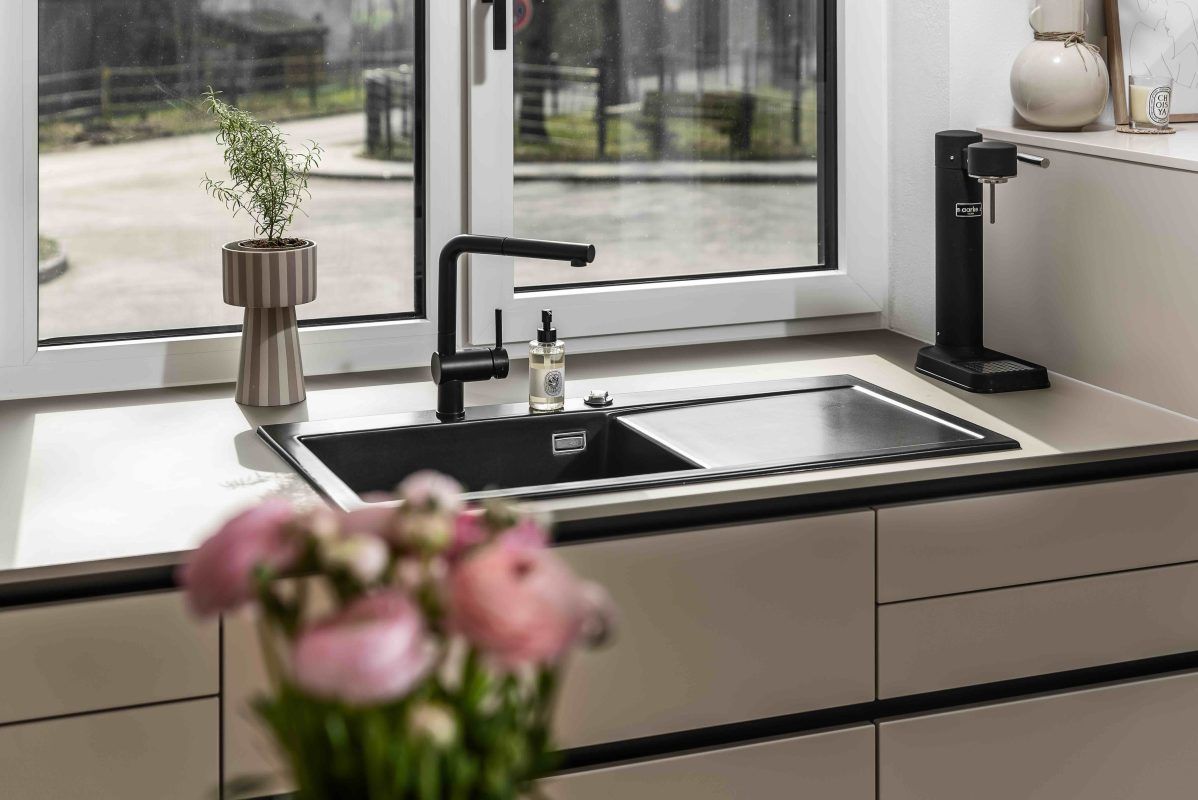 Moderne Küchenspüle neben großen Fenstern, mit einem schwarzen Wasserhahn und einer Blumenvase auf der Arbeitsplatte.