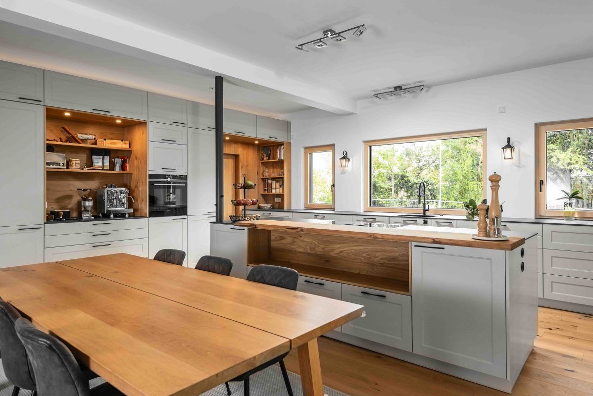Eine moderne, geräumige Küche mit hellgrauen Schränken, Oberflächen aus Naturholz, einem großen Esstisch und hellen Fenstern.
