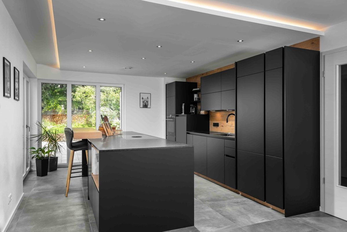 Eine moderne Küche zeichnet sich durch mattschwarze Schränke, eine Kochinsel mit Holzsitzgelegenheiten und große Fenster mit Blick ins Grüne aus.
