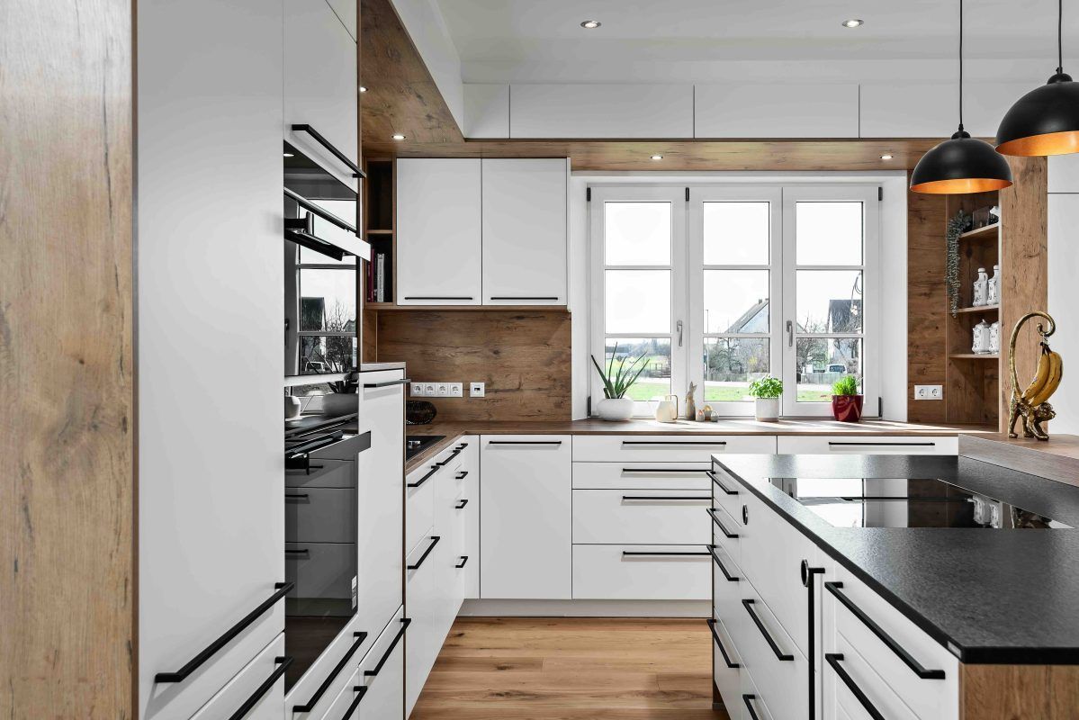 Moderne Küche mit weißen Schränken, Holzelementen, schwarzen Beschlägen und einer Kücheninsel mit Blick durch ein Fenster.