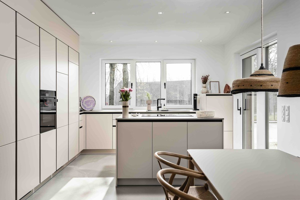 Eine moderne, minimalistische Küche mit hellbeigen Schränken, einer Kochinsel und weißen Wänden, durchflutet von natürlichem Licht.