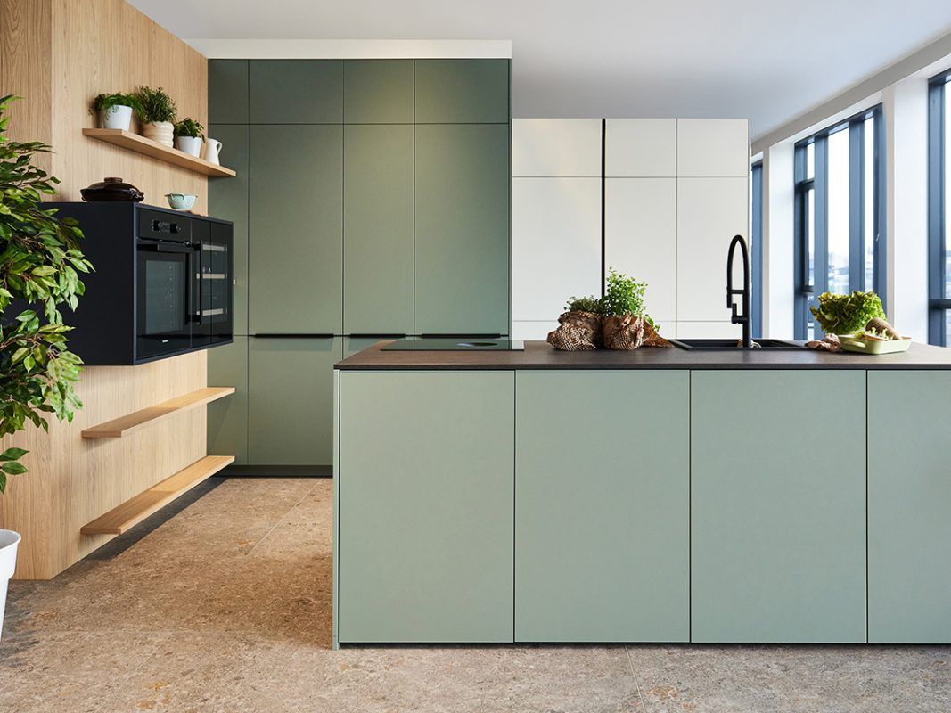 Moderne Küche mit salbeigrünen Schränken, Holzregalen und einem schwarzen Backofen neben einer großen Kücheninsel.