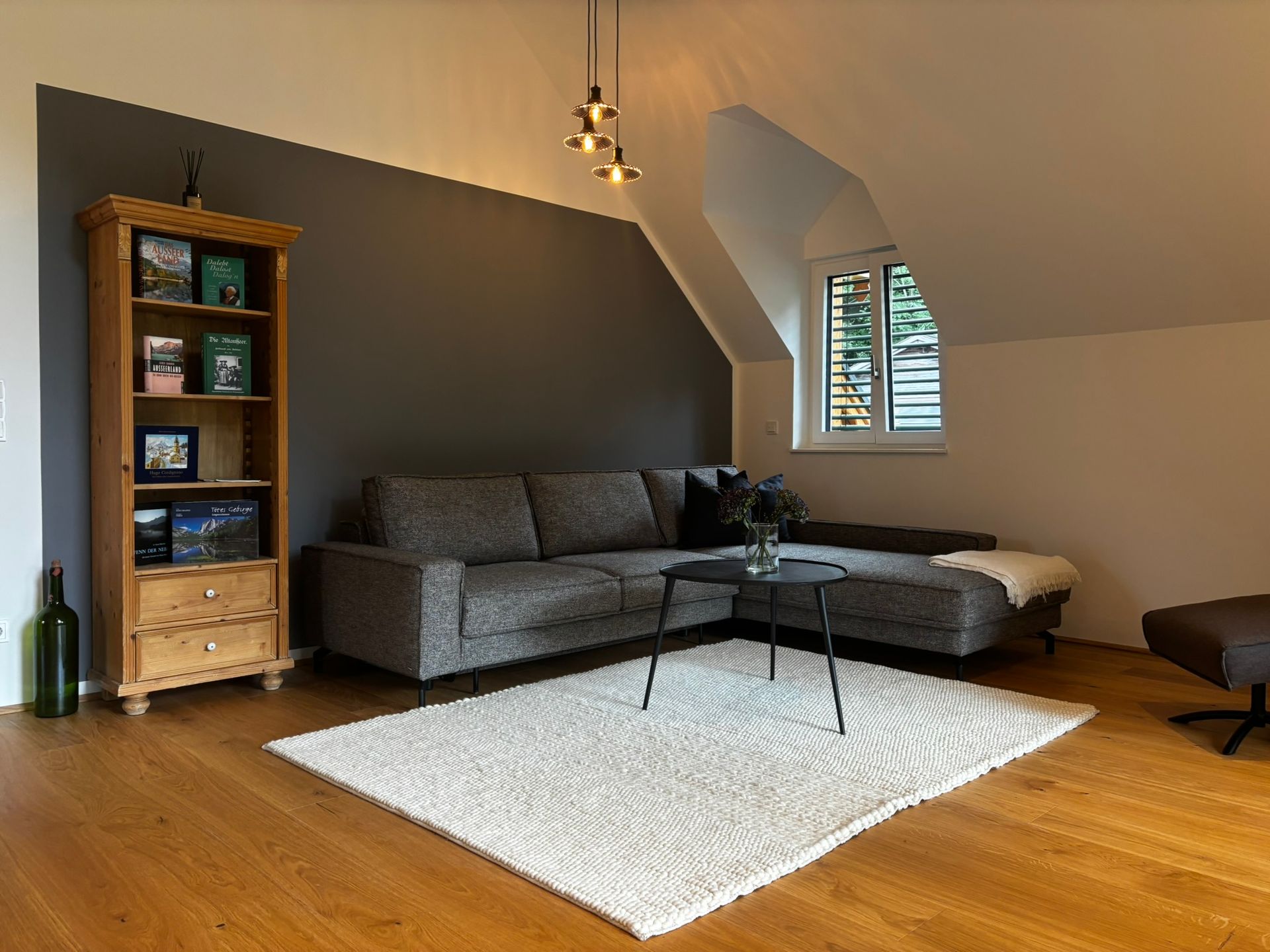 In einem Zimmer mit Holzboden, einer dunklen Akzentwand und einem hölzernen Bücherregal steht ein graues Ecksofa auf einem weißen Teppich.