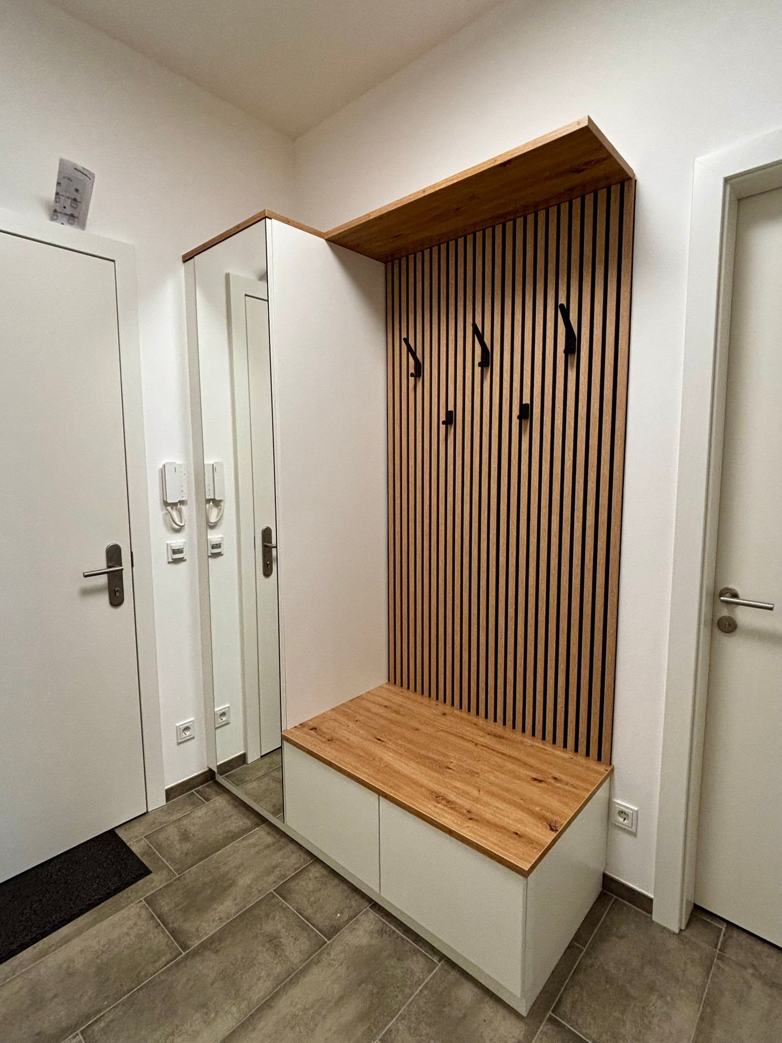 Ein moderner Eingangsbereich mit Holzlamellenwand, Kleiderhaken, einer Sitzbank und einem Ganzkörperspiegel.