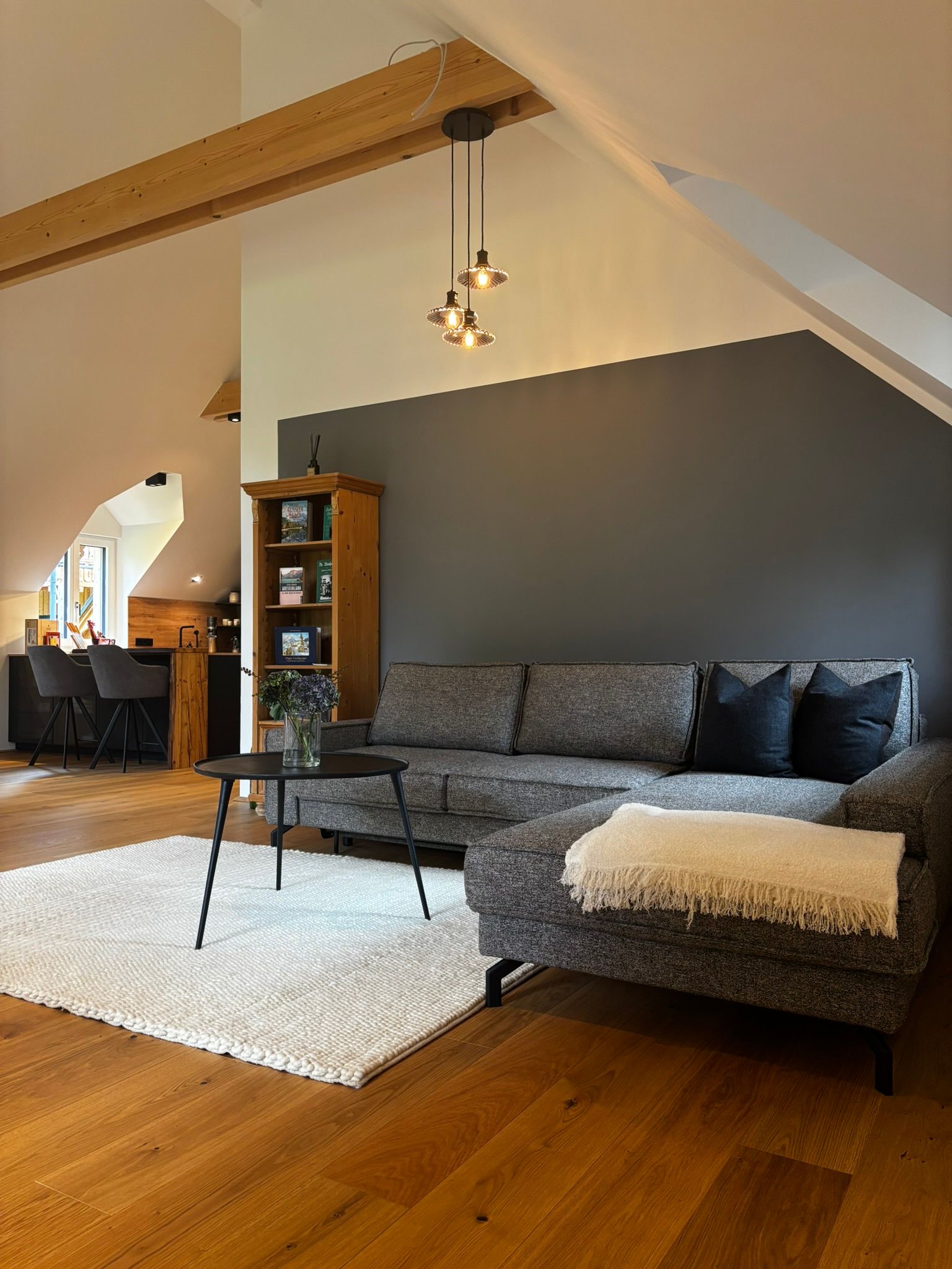 Ein gemütlicher Wohnbereich mit grauem Ecksofa, weißem Teppich, Holzregal und Pendelleuchten unter einer schrägen Decke.