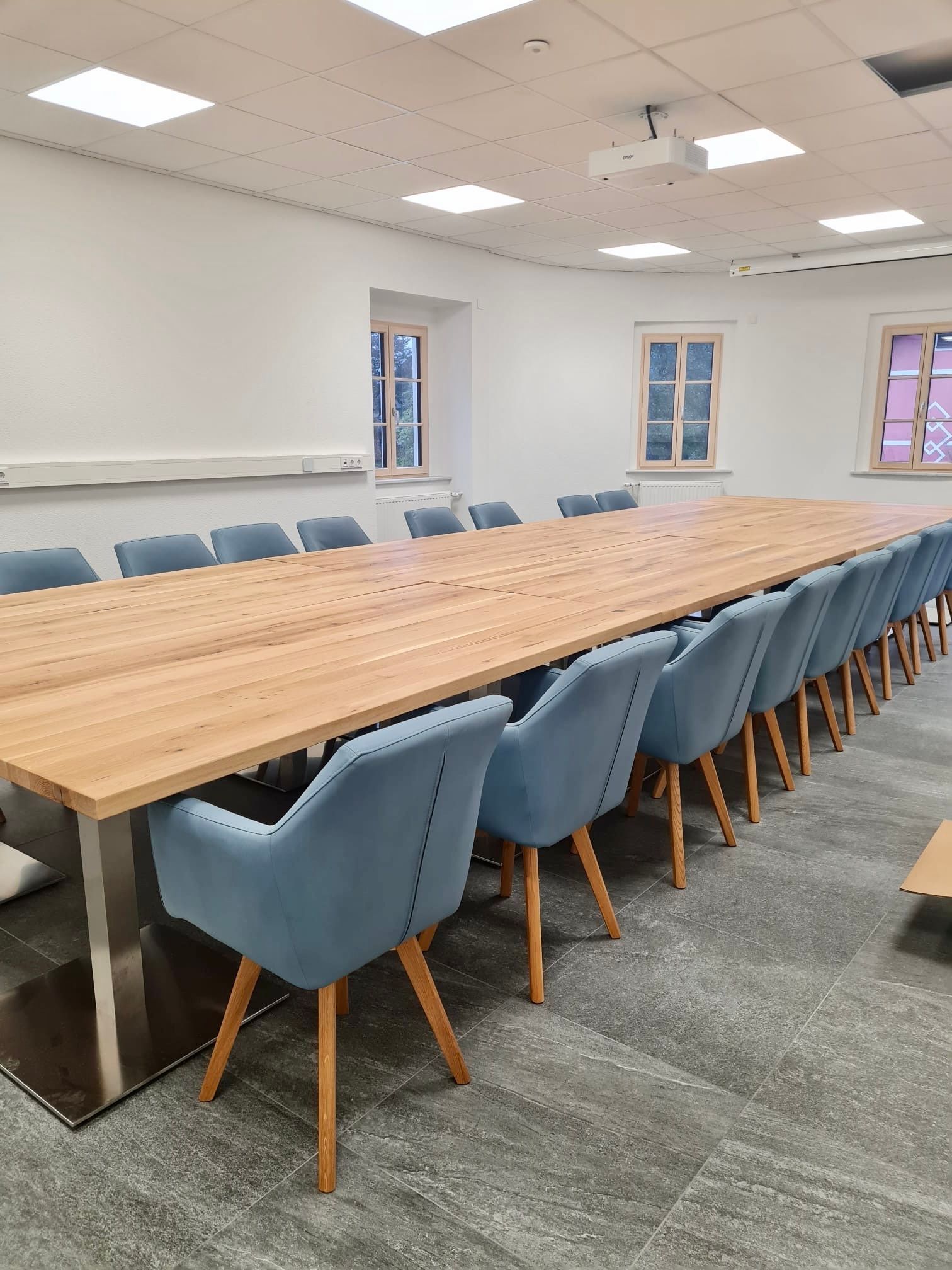 Ein langer Konferenztisch aus Holz, umgeben von blau gepolsterten Stühlen, in einem hellen, modernen Raum mit drei Fenstern.