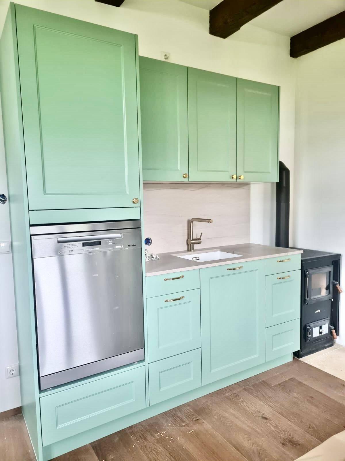 Eine Küche mit salbeigrünen Schränken, einem Geschirrspüler aus Edelstahl, einer Spüle und einem schwarzen Holzofen.