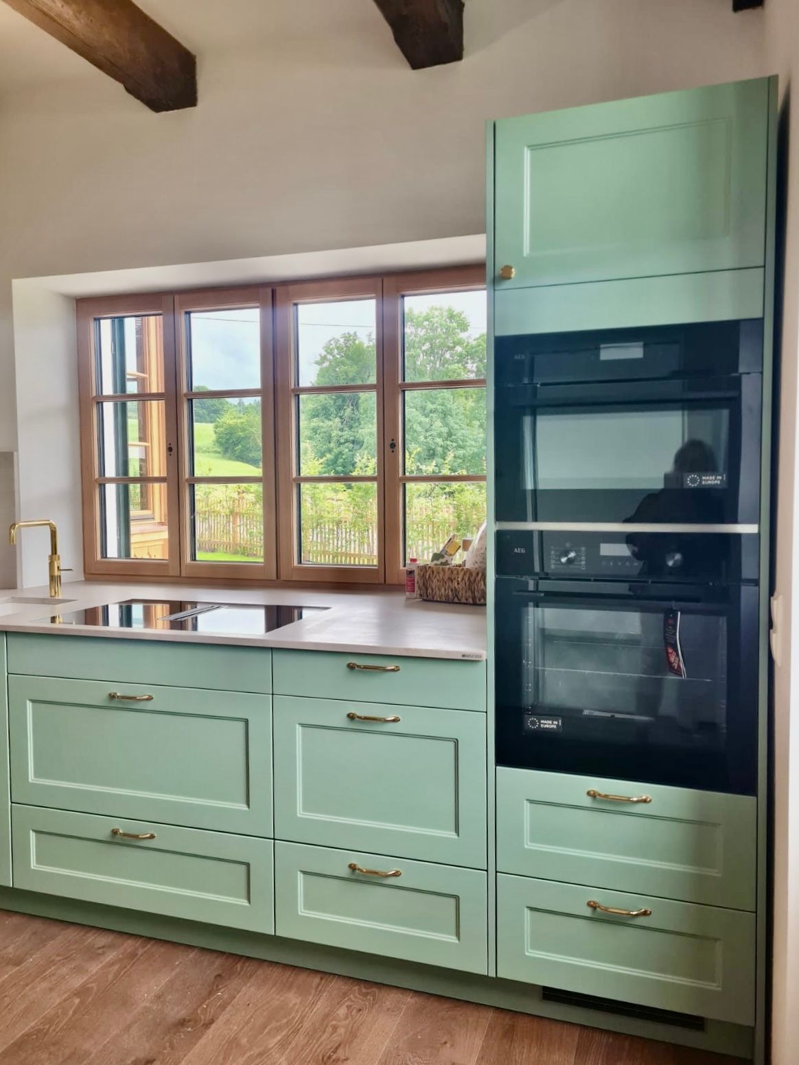 Salbeigrüne Küchenschränke mit Messinggriffen, ein eingebauter Doppelbackofen und ein Fenster mit Blick auf eine grasbewachsene ländliche Landschaft.