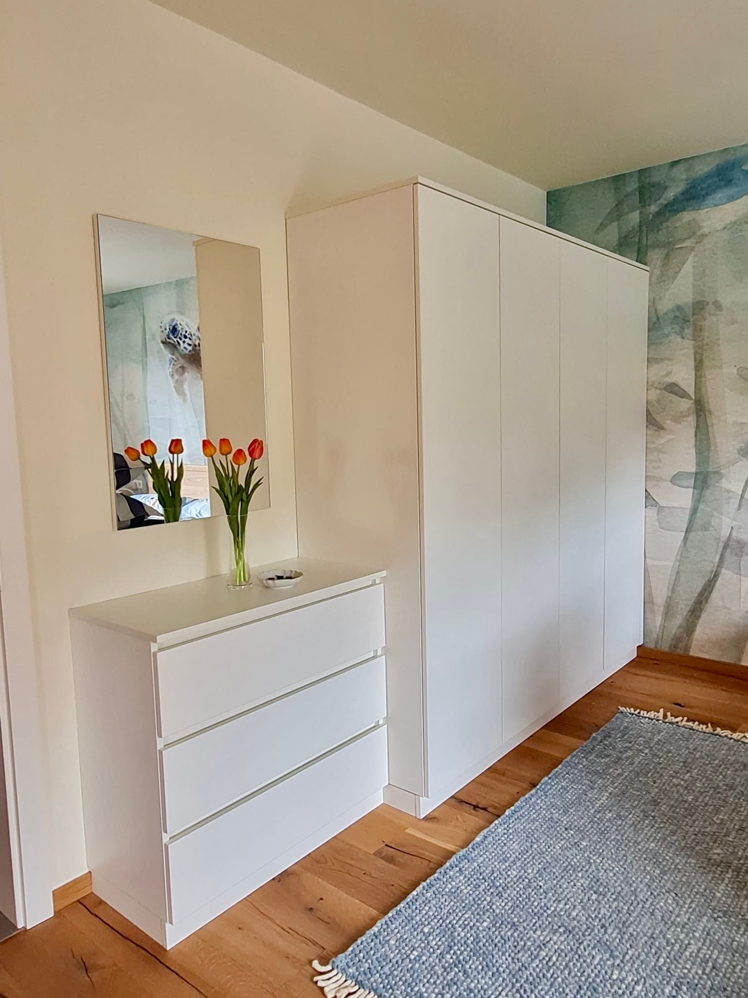 In einem hellen Zimmer stehen eine weiße Kommode und ein hoher Kleiderschrank auf einem Hartholzboden neben einem Spiegel mit Tulpen und einem blauen Teppich.