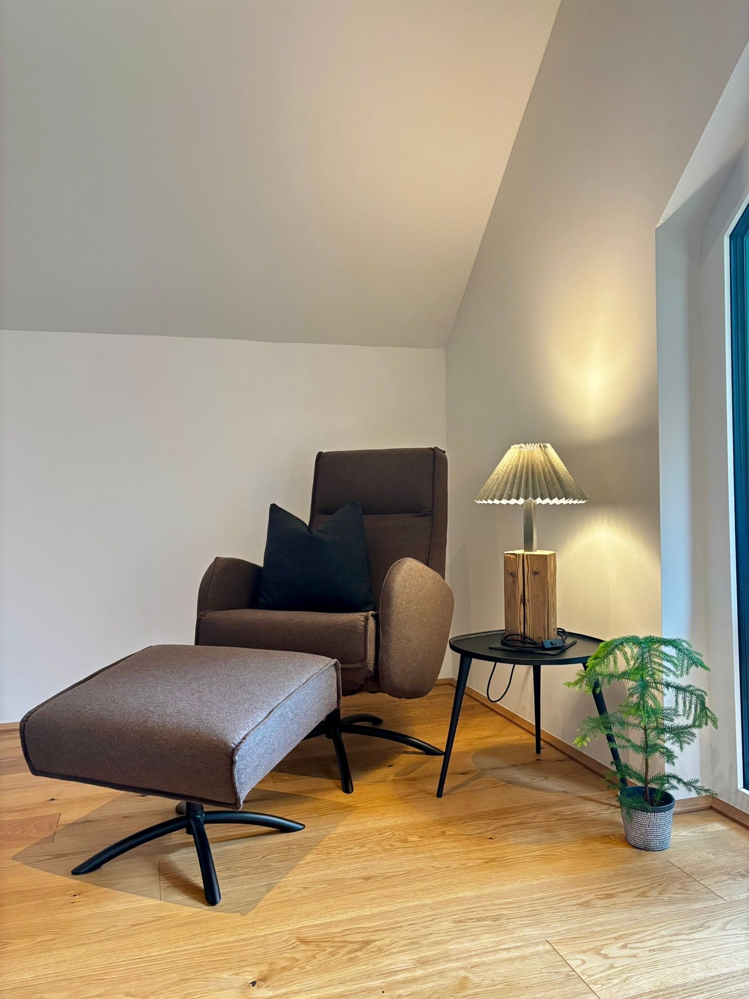 Ein brauner Sessel mit Hocker, eine Lampe auf einem kleinen runden Tisch und eine kleine Topfpflanze auf einem Holzboden.