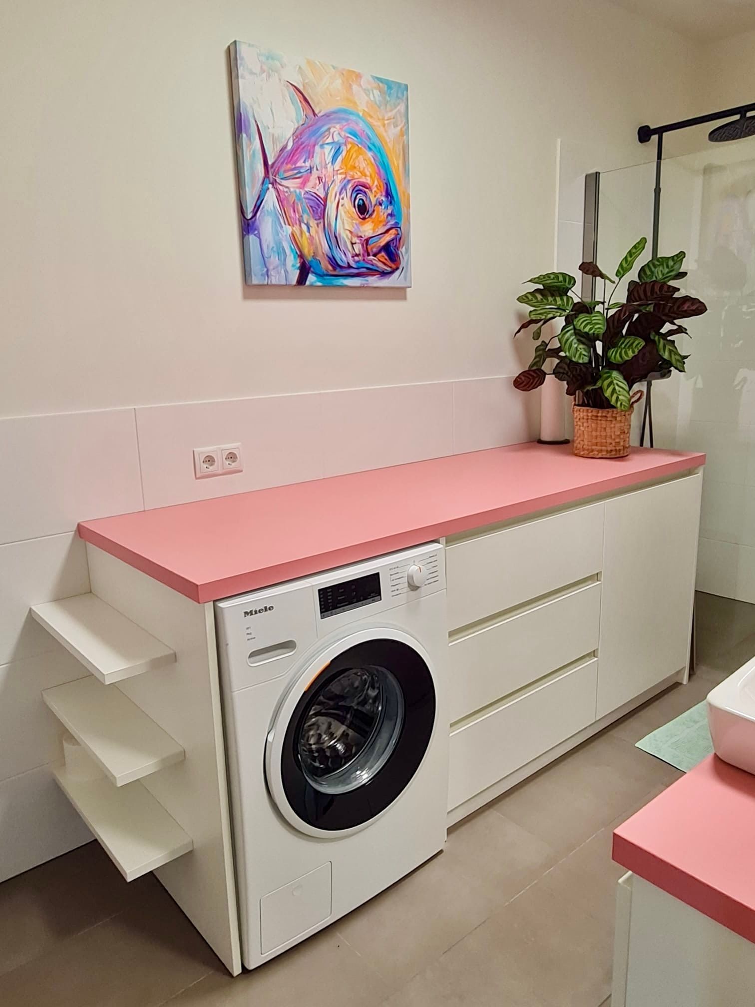Der Waschraum ist mit weißen Schränken, einer rosa Arbeitsplatte, einer weißen Waschmaschine, Wandregalen und einem farbenfrohen Fischbild ausgestattet.