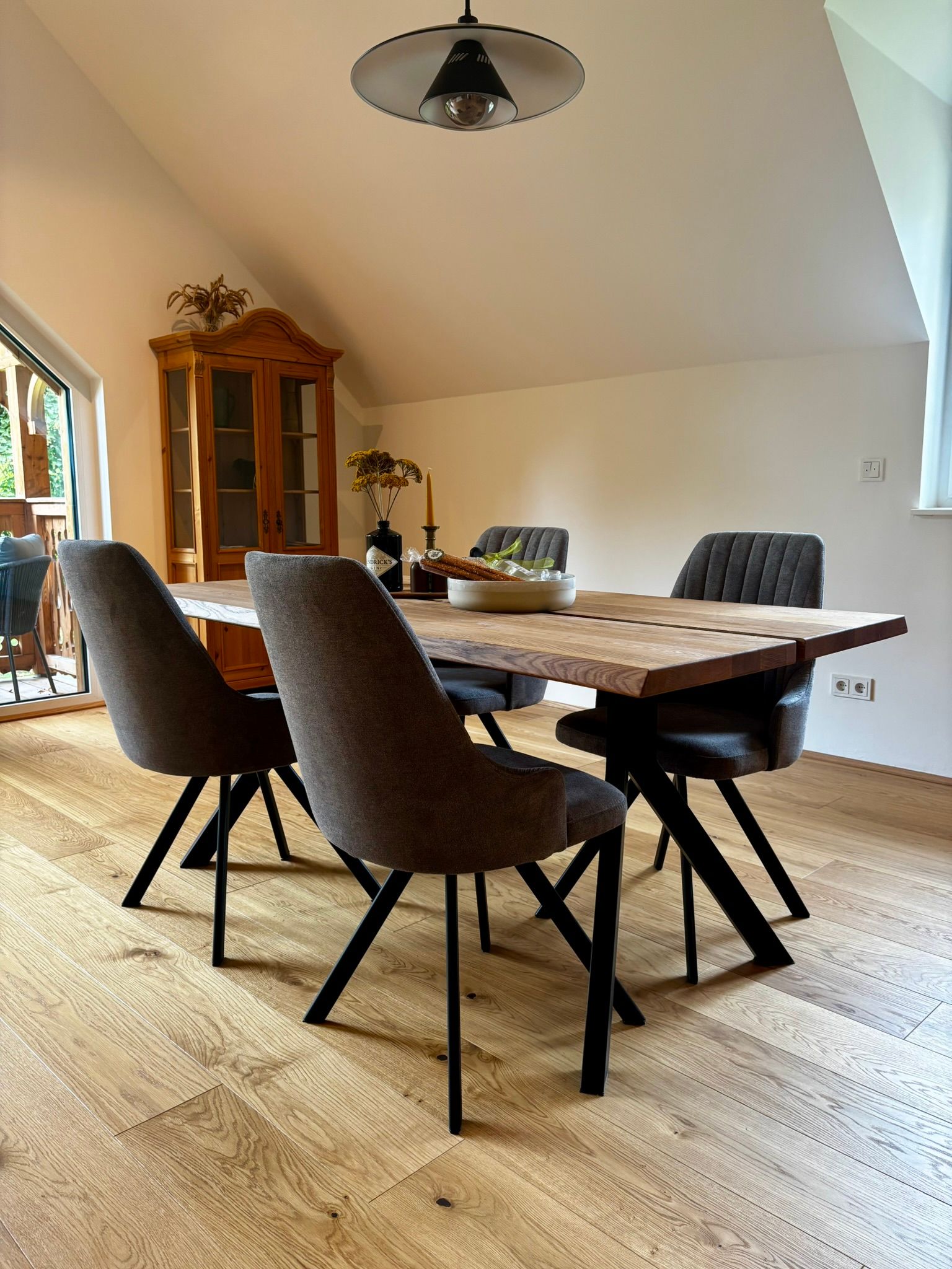 Essbereich mit einem großen Holztisch, grau gepolsterten Stühlen und einer Holzvitrine auf hellem Holzboden.