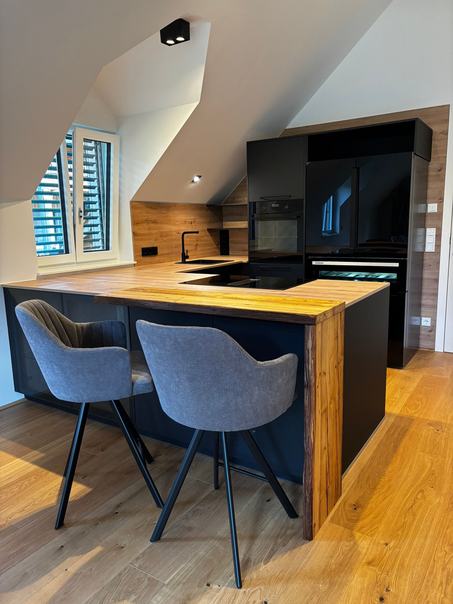 Eine moderne Küche mit einer Kücheninsel mit Holzplatte, zwei grauen Drehstühlen und schwarzen Schränken in einem Dachzimmer mit Fenster.
