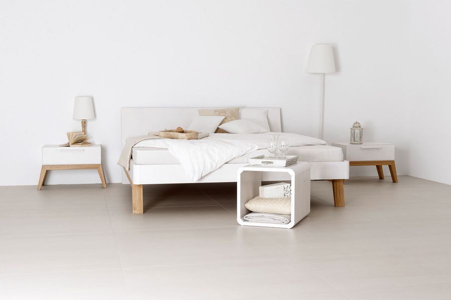 Ein minimalistisches Schlafzimmer mit einem weißen Bett, zwei Nachttischen mit Holzgestell, einer Nachttischlampe, einer Stehlampe und einem quadratischen Beistelltisch.
