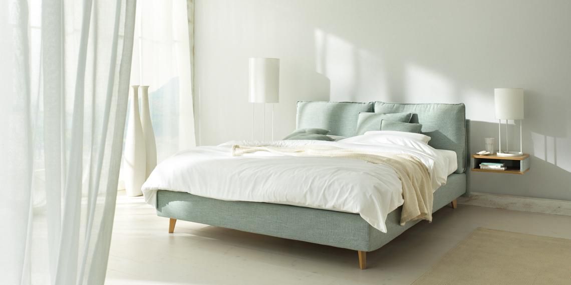 Ein hellgrünes Polsterbett mit weißer Bettwäsche, neben einem schwebenden Nachttisch und hohen Lampen in einem hellen Zimmer.