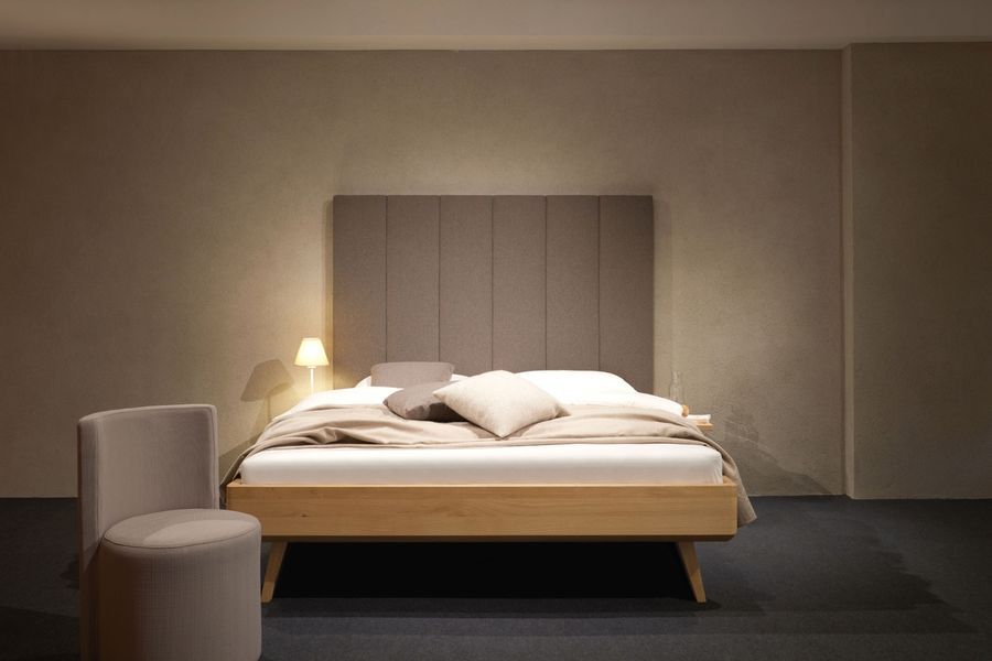 Ein modernes Bett mit hohem, vertikal gepaneeltem Kopfteil in einem minimalistischen Zimmer, dazu ein kleiner Hocker und eine warme Nachttischlampe.