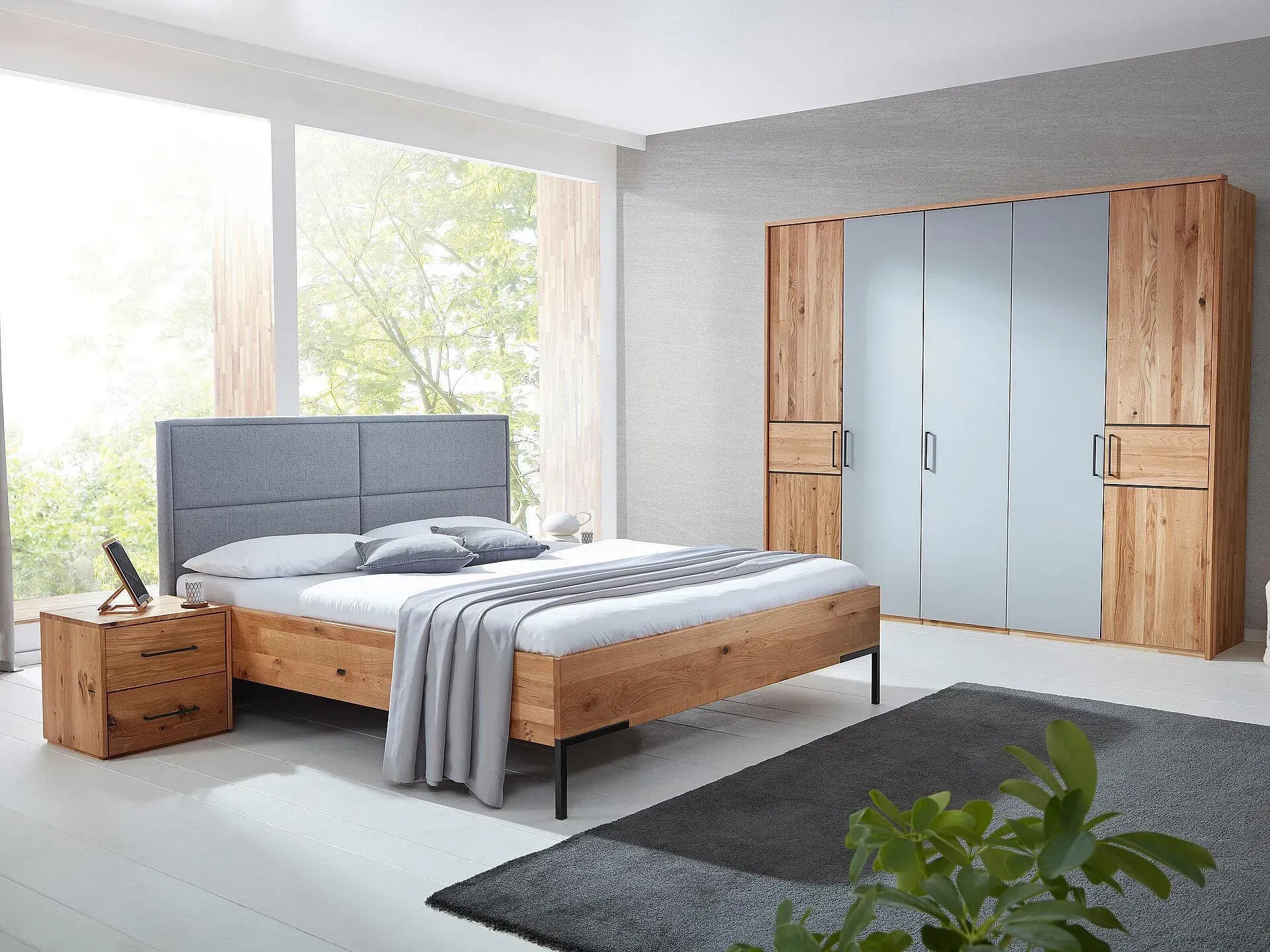 Modernes Schlafzimmer mit grauem Polsterbett, Nachttisch aus Holz, großem Kleiderschrank und hellem Fenster.