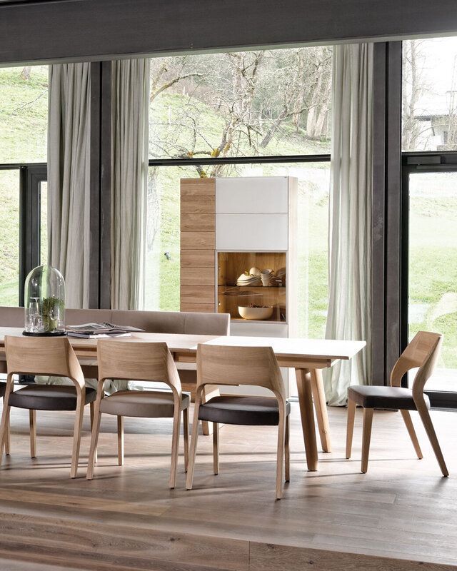 Ein Esszimmer mit einem langen Holztisch, vier modernen hellen Holzstühlen und einem hohen Schrank vor großen Fenstern.
