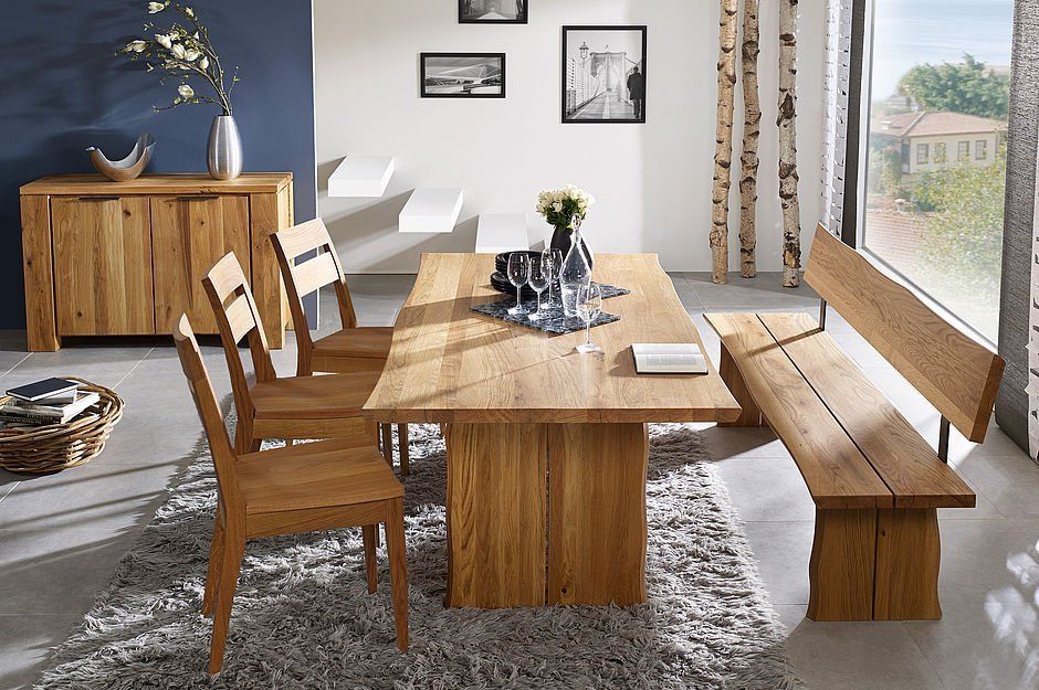 Eine Essgruppe aus Holz mit Tisch, drei Stühlen und einer Bank auf einem grauen Teppich in einem modernen Zimmer mit einer blauen Akzentwand.