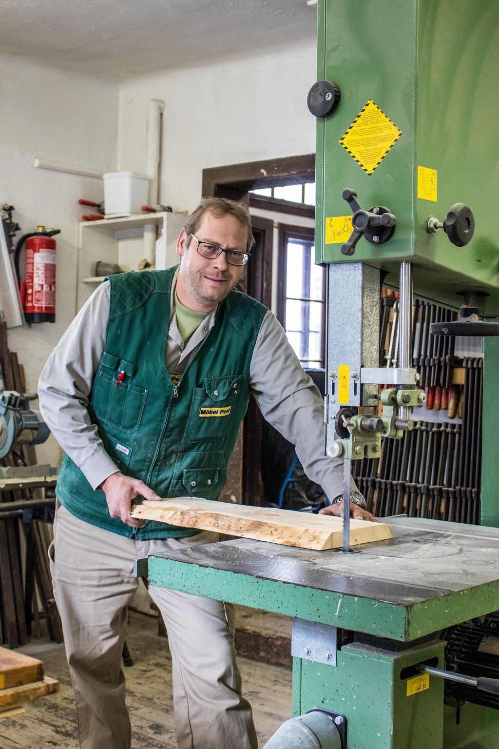 Ein Handwerker mit Schutzbrille und grüner Weste schneidet in einer Werkstatt mit einer Bandsäge ein Stück Holz zu.
