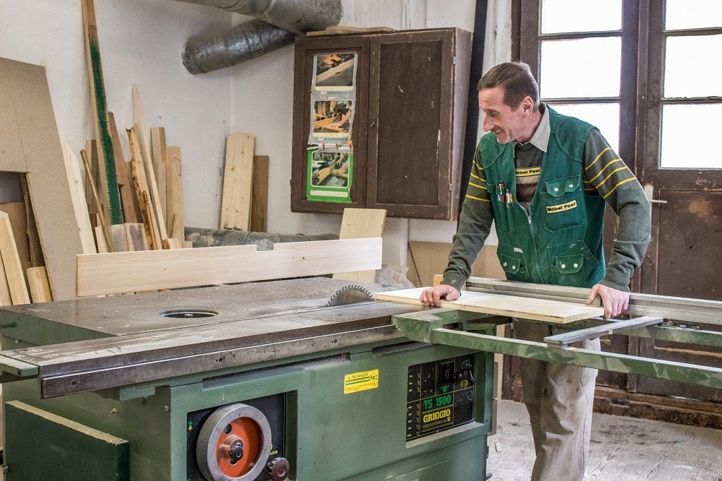 Eine Person in einer grünen Weste bedient in einer Holzwerkstatt eine Schiebetischkreissäge und führt ein Holzbrett zum Sägeblatt.