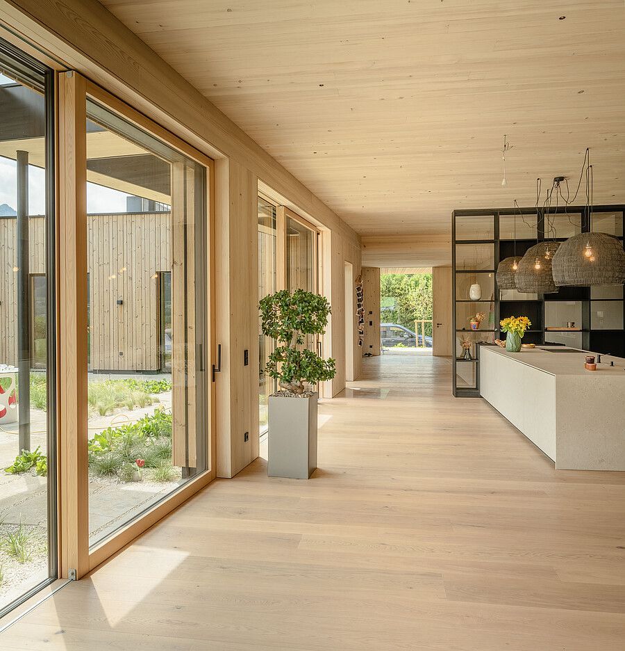 Ein minimalistischer, hellholzgetäfelter Flur mit großen Glastüren, die sich zu einem Innenhof öffnen, und einer modernen Küche auf der rechten Seite.