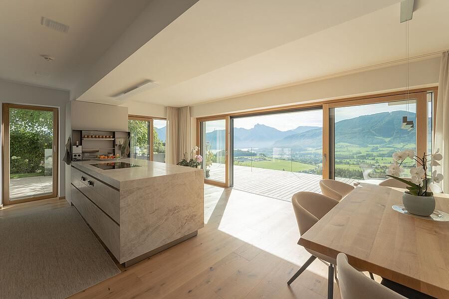 Eine moderne Küche mit Essbereich, einer Kücheninsel aus Stein und einem Holztisch, die sich zu einer Terrasse mit Bergblick öffnet.