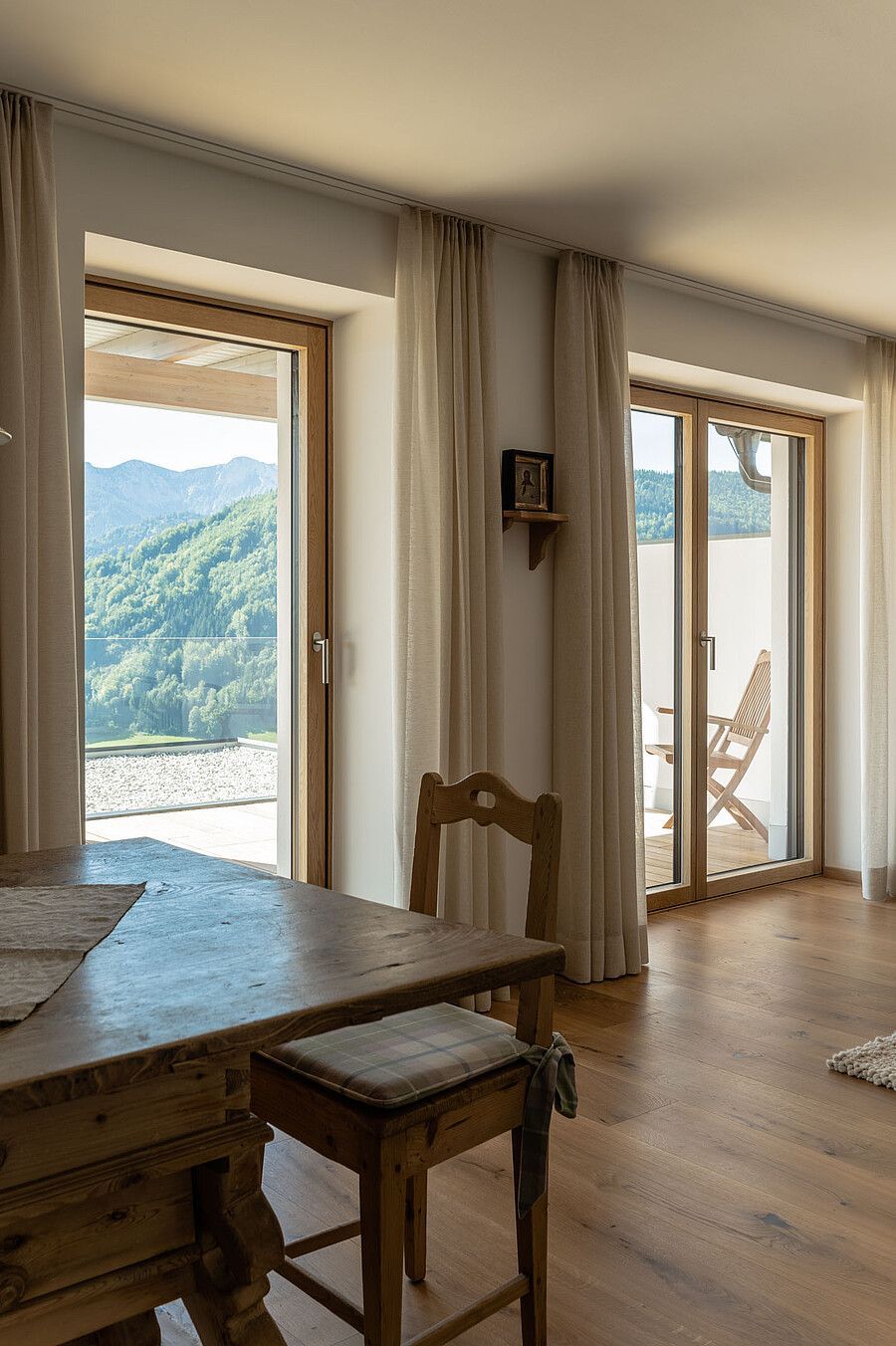 In einem hellen Raum mit zwei bodentiefen Glastüren, die den Blick auf eine Berglandschaft freigeben, stehen ein Holztisch und ein Hocker.