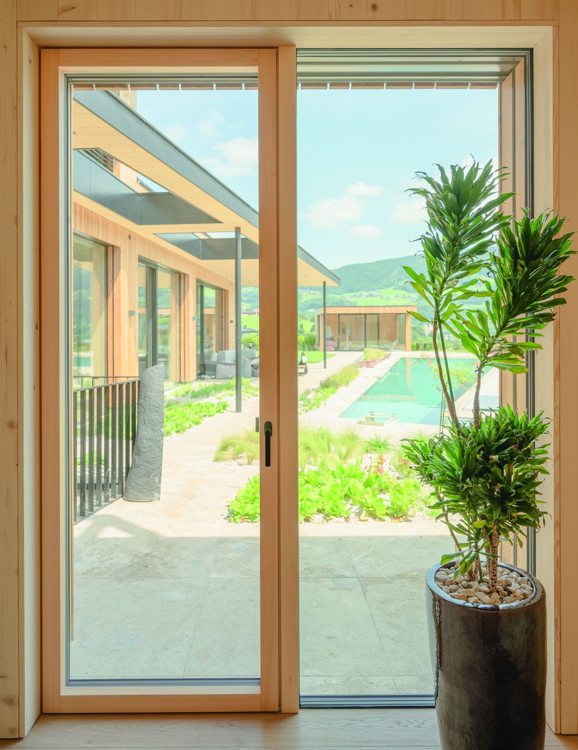 Der Blick durch eine holzgerahmte Glastür geht auf eine Terrasse, einen Swimmingpool und eine Hügellandschaft mit einer Topfpflanze über.