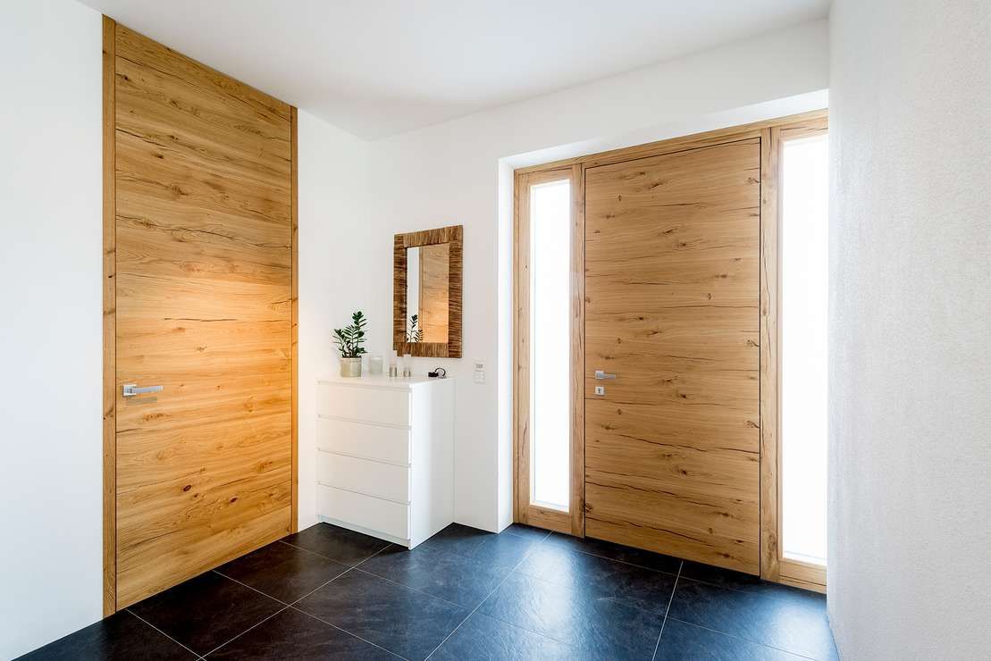 Ein heller Eingangsbereich mit dunklen Fliesenböden, einer Holztür, einem passenden Seiteneingang mit Glastür und einer kleinen weißen Truhe.